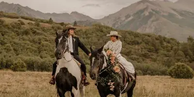 A bride and groom in western-style wedding attire riding horses side-by-side in a grassy field with mountains in the background.