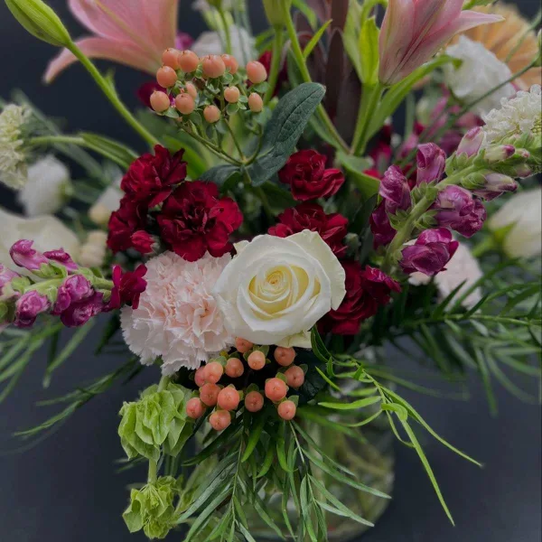 A close-up of a mixed floral arrangement in a glass vase featuring pink lilies, a white rose, red carnations, and greenery.