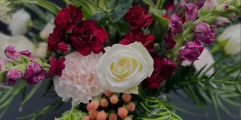 A close-up of a mixed floral arrangement in a glass vase featuring pink lilies, a white rose, red carnations, and greenery.