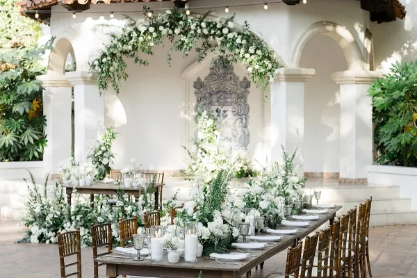 A wedding venue featuring a long wooden table set with white floral centerpieces, candles, and glassware, situated under a white archway decorated with hanging greenery and white flowers.