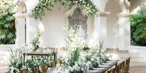 A wedding venue featuring a long wooden table set with white floral centerpieces, candles, and glassware, situated under a white archway decorated with hanging greenery and white flowers.