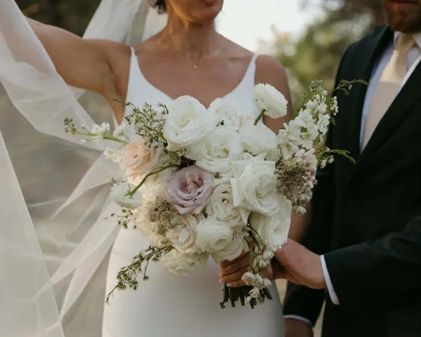 A bride in a white dress holding a large bouquet of white and pale pink flowers, including roses and ranunculus, with a groom in a dark suit standing beside her.