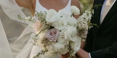 A bride in a white dress holding a large bouquet of white and pale pink flowers, including roses and ranunculus, with a groom in a dark suit standing beside her.