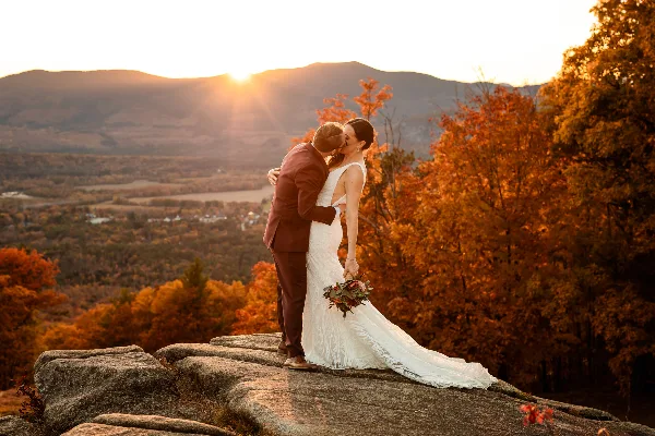 A bride and groom kissing on a rocky outcrop with a scenic mountain and autumn foliage background at sunset.