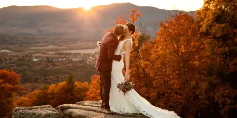 A bride and groom kissing on a rocky outcrop with a scenic mountain and autumn foliage background at sunset.
