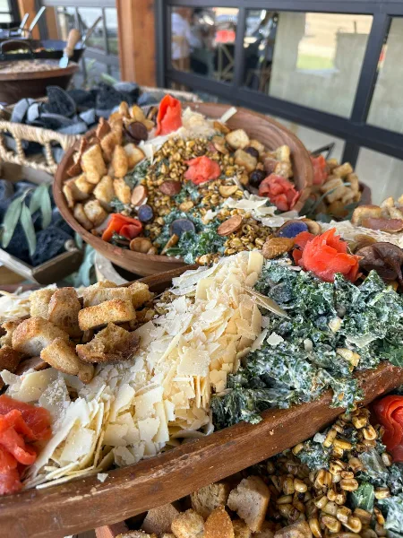 A close-up of a large wooden platter filled with a salad, croutons, shaved cheese, and roasted vegetables.