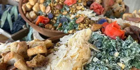 A close-up of a large wooden platter filled with a salad, croutons, shaved cheese, and roasted vegetables.