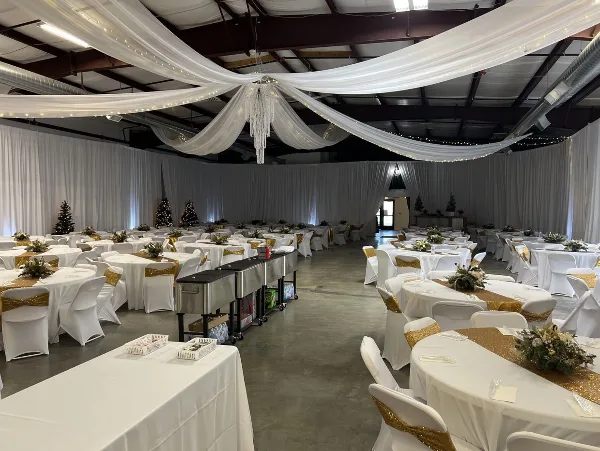 A spacious indoor wedding venue with round tables, white and gold decor, and ceiling draping.