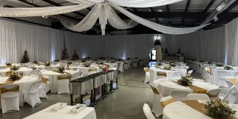 A spacious indoor wedding venue with round tables, white and gold decor, and ceiling draping.