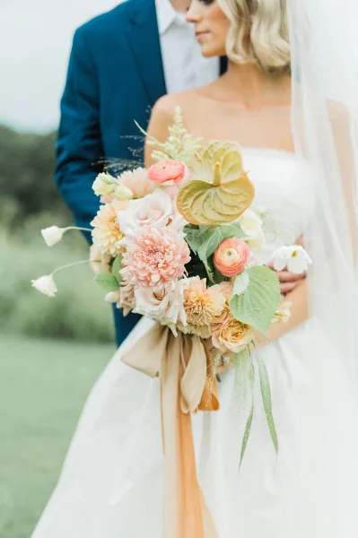 A bride in a white gown and veil holding a large floral bouquet with a groom in a blue suit standing behind her.