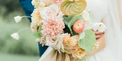 A bride in a white gown and veil holding a large floral bouquet with a groom in a blue suit standing behind her.