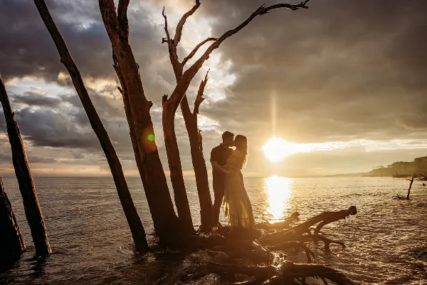 A couple embracing in a romantic outdoor setting near water with a dramatic sunset sky.