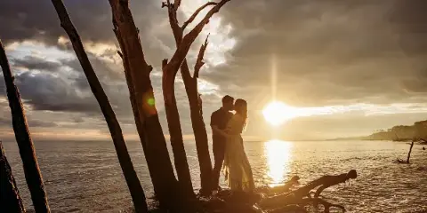 A couple embracing in a romantic outdoor setting near water with a dramatic sunset sky.