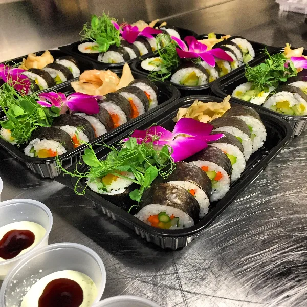 Multiple black catering trays filled with sushi rolls, garnished with pink orchids and green microgreens, sitting on a metal kitchen counter.