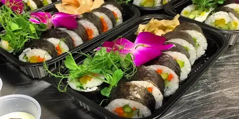 Multiple black catering trays filled with sushi rolls, garnished with pink orchids and green microgreens, sitting on a metal kitchen counter.