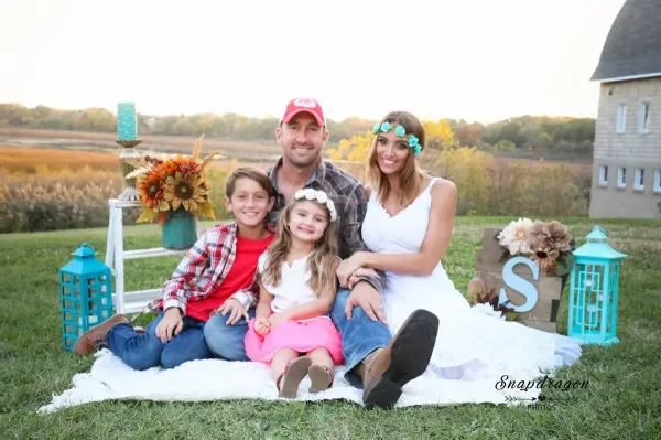 A bride, groom, and two children sitting on a white blanket in a grassy field with a historic stone building in the background.