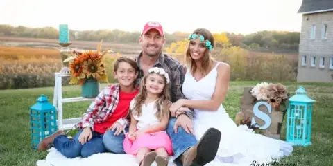 A bride, groom, and two children sitting on a white blanket in a grassy field with a historic stone building in the background.