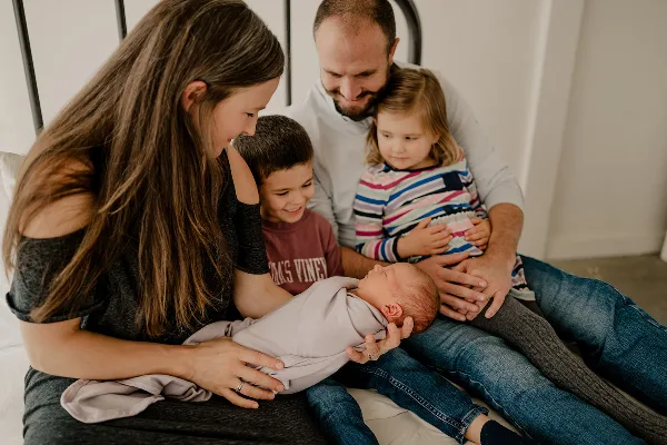 A family of five, including a mother holding a newborn, a father holding a toddler, and a young boy, posing together on a bed.