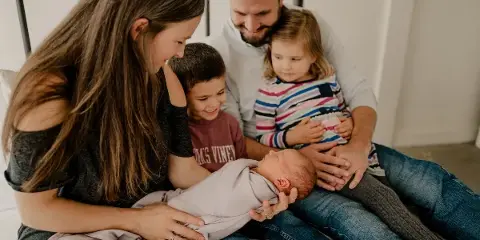 A family of five, including a mother holding a newborn, a father holding a toddler, and a young boy, posing together on a bed.