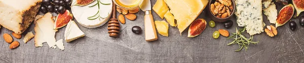 A flat lay of gourmet catering ingredients featuring various cheeses, fresh figs, almonds, and a honey dipper on a dark surface.