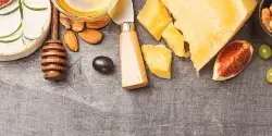 A flat lay of gourmet catering ingredients featuring various cheeses, fresh figs, almonds, and a honey dipper on a dark surface.