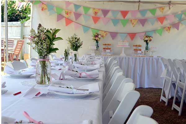 A long banquet table set with white tablecloths, plates, and floral centerpieces under a tent decorated with colorful bunting.