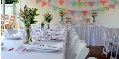 A long banquet table set with white tablecloths, plates, and floral centerpieces under a tent decorated with colorful bunting.