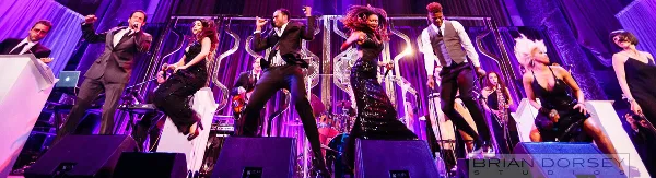 A live band performing on a stage with purple lighting, featuring multiple vocalists and musicians.