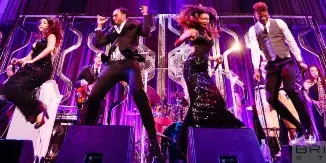 A live band performing on a stage with purple lighting, featuring multiple vocalists and musicians.