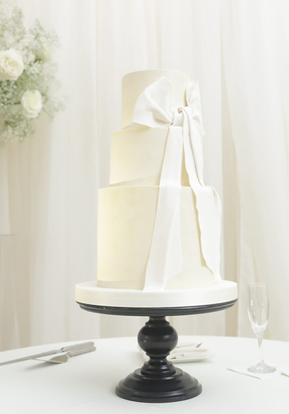 A three-tier white wedding cake with a large fabric-style bow on a black pedestal stand.