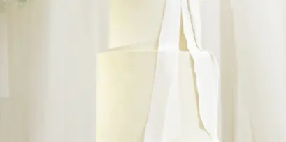 A three-tier white wedding cake with a large fabric-style bow on a black pedestal stand.