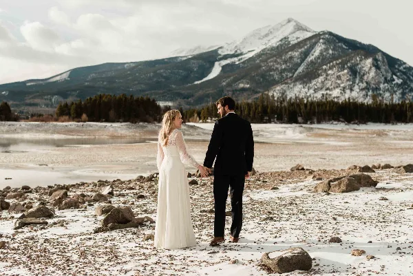 A bride and groom holding hands in a snowy landscape with mountains in the background.