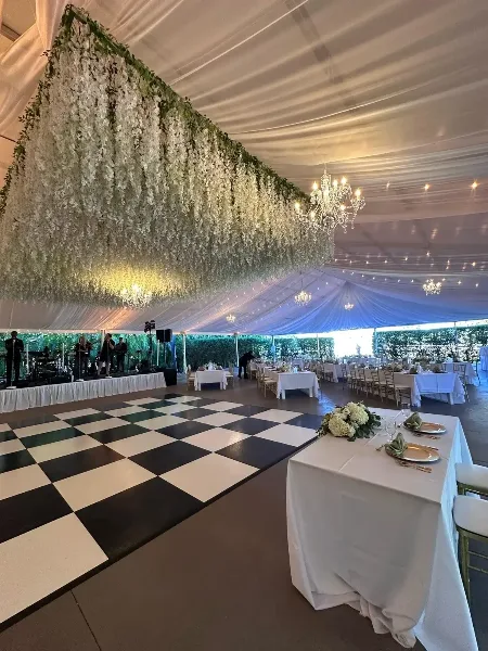 An elegant event venue featuring a large white floral wall, chandeliers, and a checkered dance floor with set dining tables.