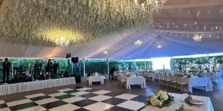 An elegant event venue featuring a large white floral wall, chandeliers, and a checkered dance floor with set dining tables.
