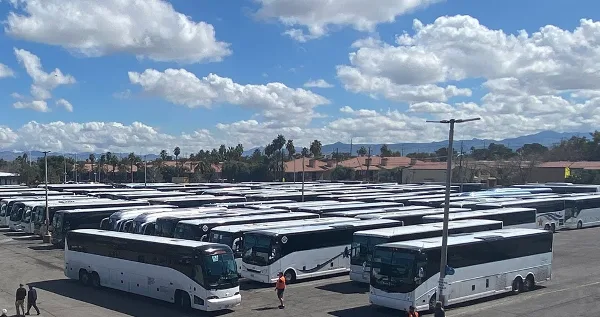 A large fleet of white charter buses parked in a lot under a blue sky with clouds.