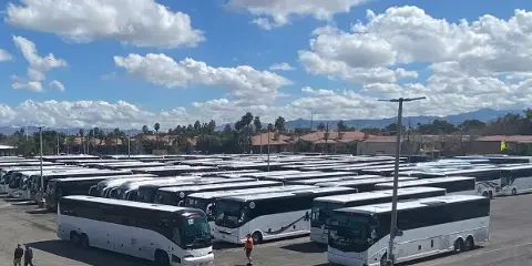 A large fleet of white charter buses parked in a lot under a blue sky with clouds.