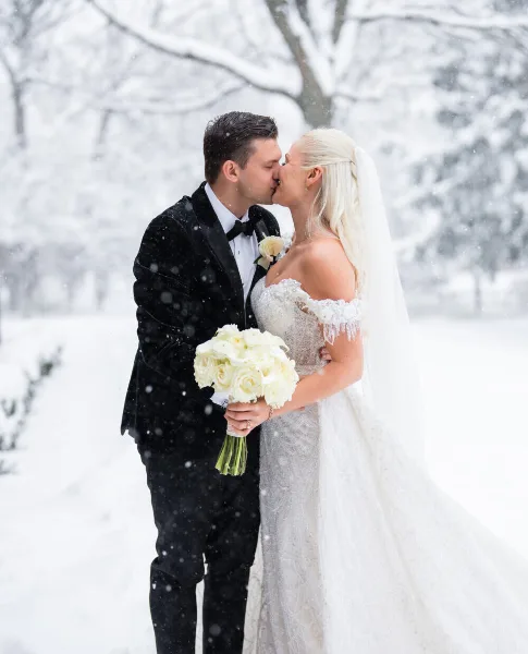 A bride and groom sharing a kiss in a snowy landscape, with the bride holding a white bouquet.