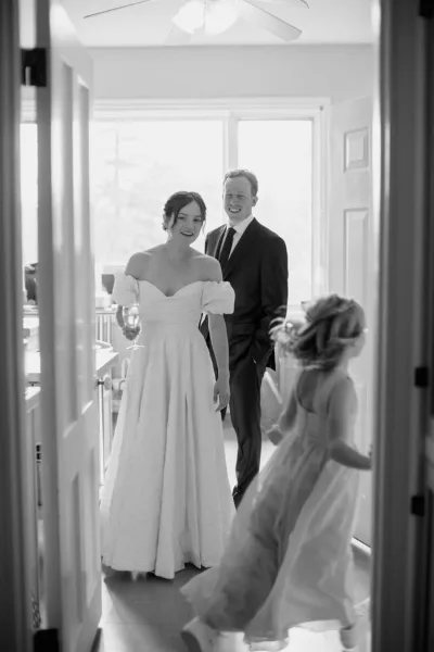 A black and white photograph of a bride and groom standing in a doorway, with a young girl in the foreground.