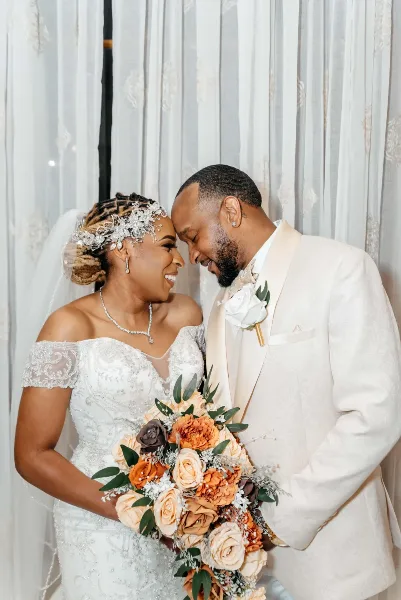 A bride in a white lace gown and a groom in a cream suit standing close together, holding a bouquet of orange and white flowers.