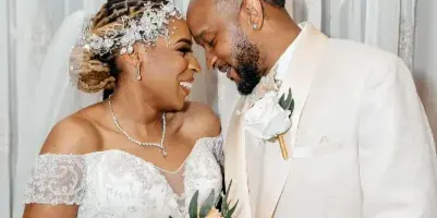 A bride in a white lace gown and a groom in a cream suit standing close together, holding a bouquet of orange and white flowers.