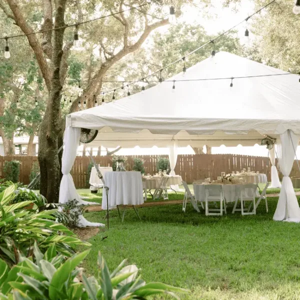 A white event tent set up on a lawn with tables, chairs, and string lights.