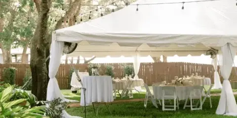 A white event tent set up on a lawn with tables, chairs, and string lights.