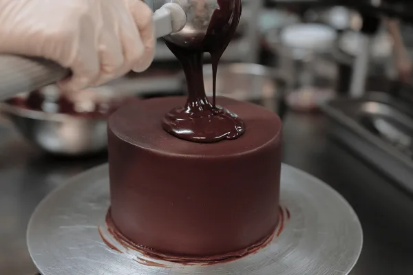 A gloved hand pouring chocolate ganache onto a round cake on a turntable.