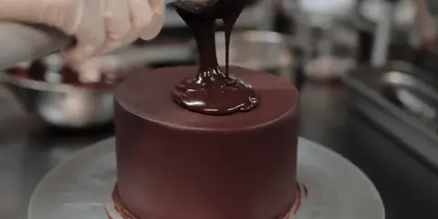 A gloved hand pouring chocolate ganache onto a round cake on a turntable.