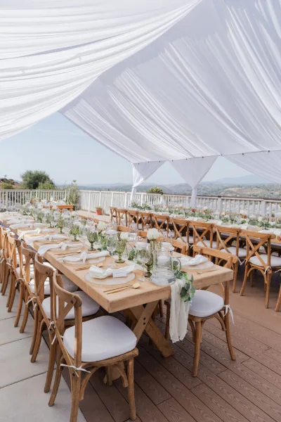 Outdoor wedding reception setup with wooden farm tables, crossback chairs, white linens, tableware, and a draped white canopy.