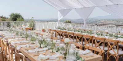 Outdoor wedding reception setup with wooden farm tables, crossback chairs, white linens, tableware, and a draped white canopy.