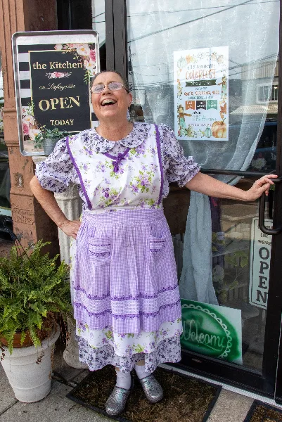 A smiling woman in a floral dress and apron stands in front of the Kitchen on Lafayette storefront with an open sign and window posters.
