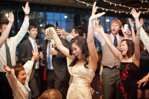 A bride and guests dancing at a wedding reception with a DJ in the background.
