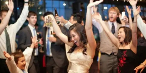 A bride and guests dancing at a wedding reception with a DJ in the background.