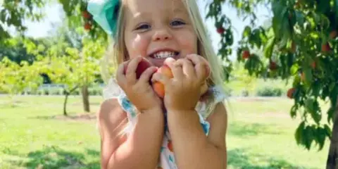 A young girl smiling while holding a peach in an orchard.